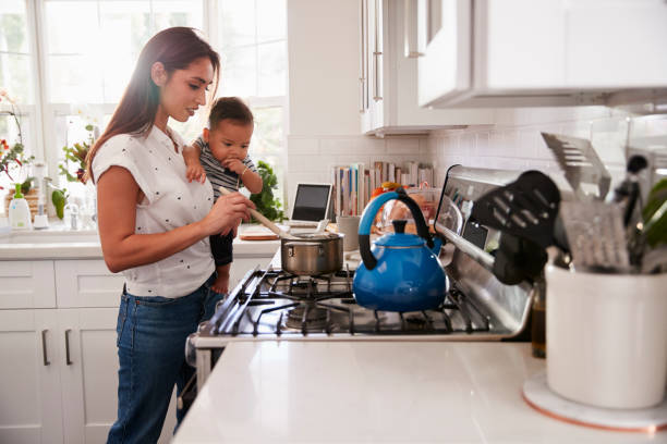 Multitasking mum holding her young baby while she makes food at the hob in her kitchen, side view