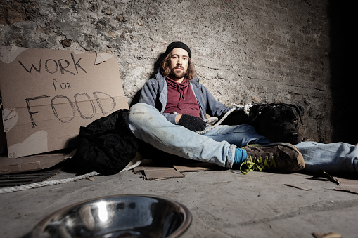 Portrait of young man wearing dirty rags hugging his dog, sitting on the city sidewalk with cardboard \