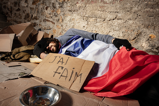 Homeless man covered with French flag, sleeping on the street next to the cardboard Jai faim sign and alms bowl