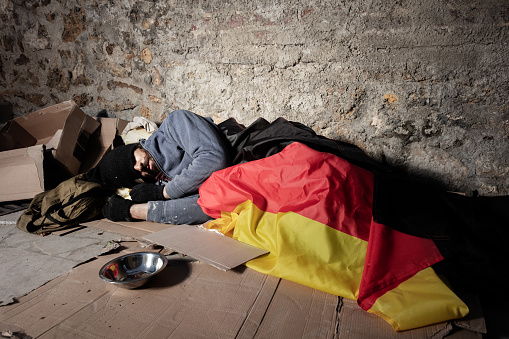 Houseless man covered with German flag, sleeping on the street next to the blanked cardboard sign and alms bowl