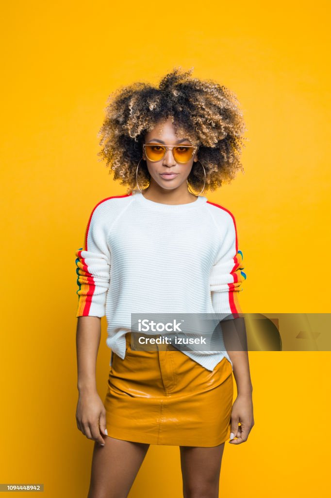 Confident, beautiful afro young woman against yellow background Confident afro young woman wearing blouse with rainbow pattern standing against yellow background, looking at camera. Studio shot. Fashion Stock Photo