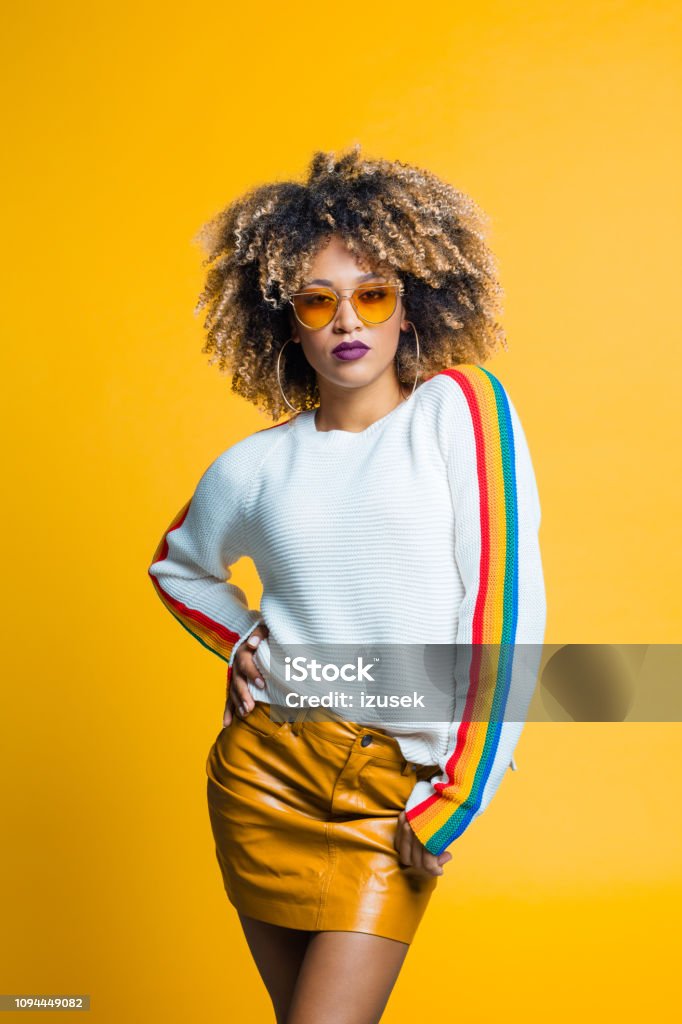Confident, beautiful afro young woman against yellow background Confident afro young woman wearing blouse with rainbow pattern standing against yellow background, looking at camera. Studio shot. Eyeglasses Stock Photo