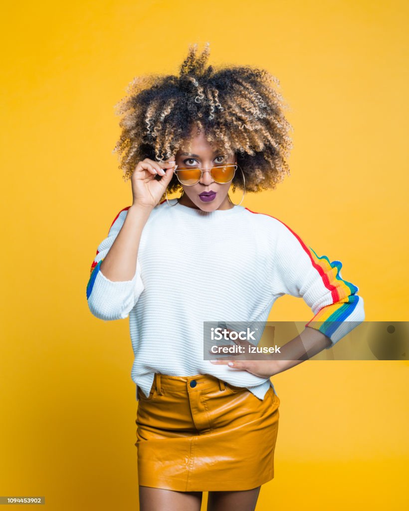 Surprised afro young woman against yellow background Surprised, beautiful afro young woman wearing blouse with rainbow pattern and sunglasses standing against yellow background. Studio shot. Fashion Stock Photo