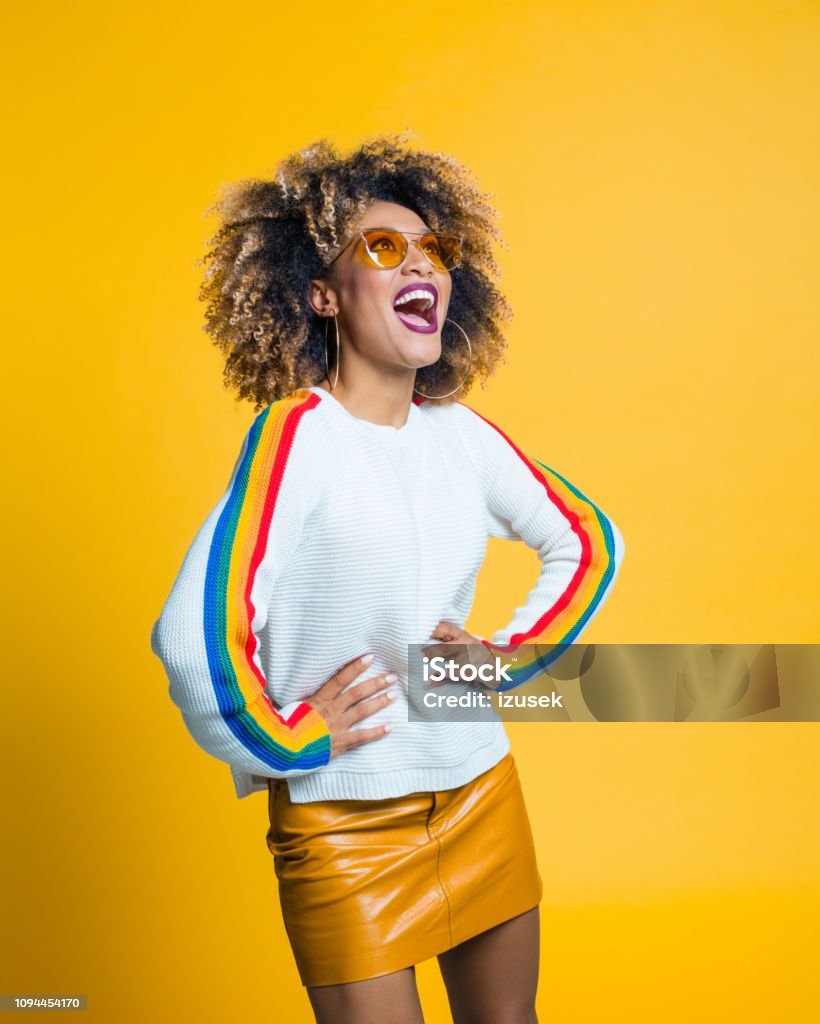Excited afro girl standing against yellow background Happy and energetic afro young woman wearing blouse with rainbow pattern laughing against yellow background. Studio shot. Women Stock Photo