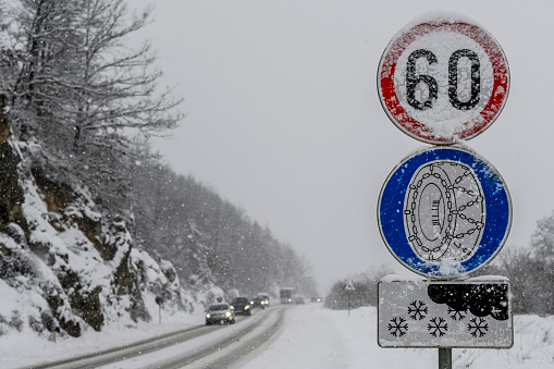 Snow chain sign with speed limit next to a winding road in wintertime. There is some traffic on the road.
