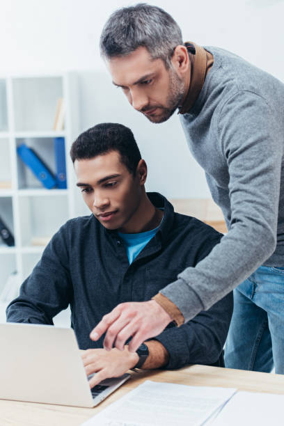 istock bearded business mentor looking at young colleague using laptop in office 1094462964