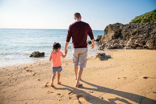 Father and young daughter walking along and exploring a beach while holding hands during a vacation