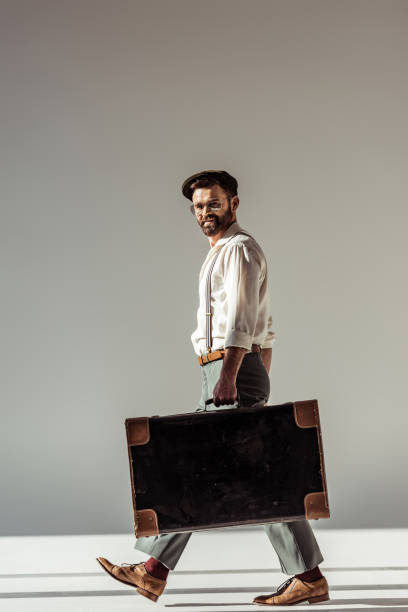 istock handsome bearded man in glasses and cap walking with retro suitcase and looking at camera on grey background 1094469900