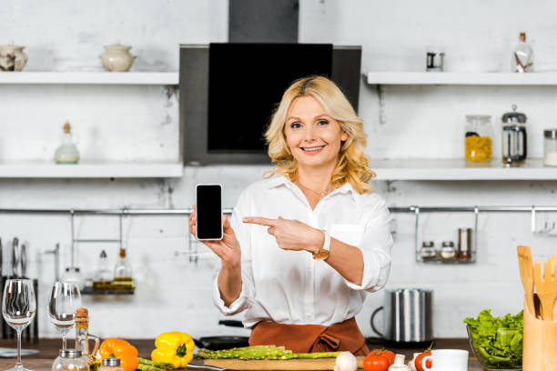 istock attractive middle aged woman pointing on smartphone with blank screen in kitchen 1094473300