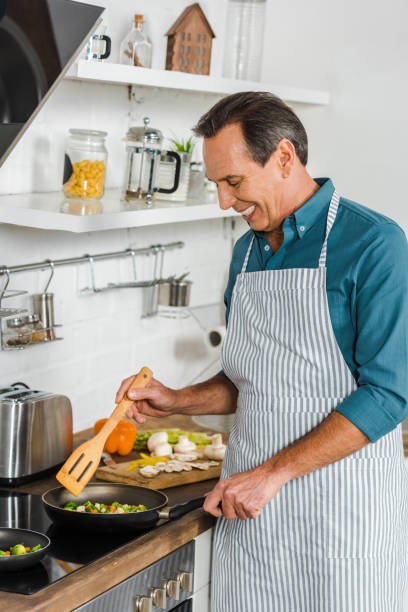 istock smiling handsome mature man cooking vegetables on frying pan in kitchen 1094473534