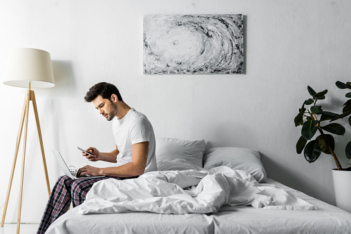 young man in pajamas using smartphone and laptop in bedroom