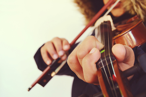 A child playing the stringed and bowed musical instrument, the violin, captured from the front. Right aligned in the horizontal shot. Empty copy space to the left.  Depth of field. The child is holding the violin bow in his hand, four fingers, and is pressing it against the strings in a beautiful pose.  There is a brown faux fur attached to the hood, hoodie of his black coat, jacket. Apt for Valentine / valentine's day, love greetings card, poster, sorry or Thank you, Propose, proposal message /  messages. No text. The bow stick is also over the child's lips.