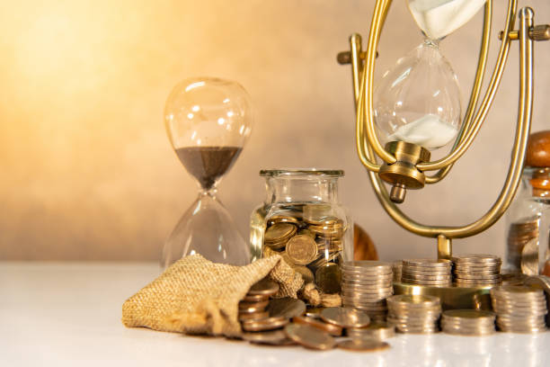 istock Sand running through the shape of hourglass with coins stack and currency jars on table. Time investment. Retirement saving concept. Urgency countdown timer for business payment deadline 1094496960