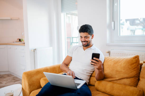 istock Young smiling african american man using laptop and mobile phone while sitting on sofa. 1094499730