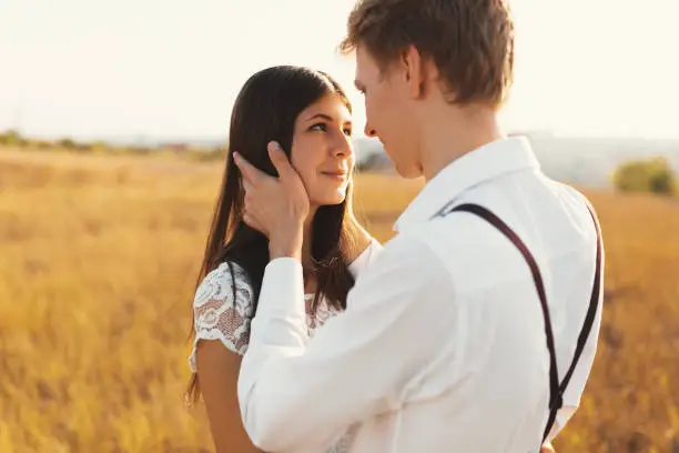 Photo of Man touching, gentle woman hair, smiling while looking into her eyes, with a beautiful landscape in background, golden grass