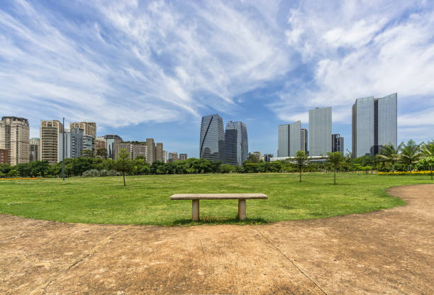 Concrete bench to sit on the grassy lawn in the park with skyline of modern buildings and mirrored on sunny day with sparse clouds stock photo