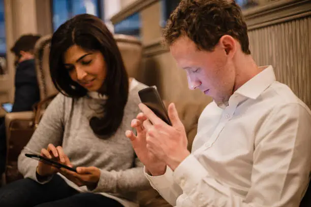 A man holding his smartphone in his hand and he is using a visually impaired mobile app to help assist him, a female can be seen sitting down next to him.