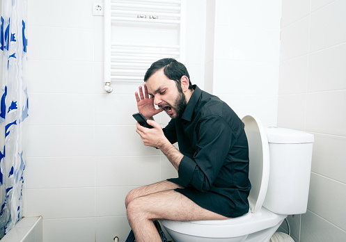 Vista lateral de un hombre frustrado gritando mientras usa un teléfono inteligente en el asiento del inodoro photo