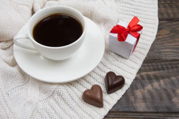 White cup with black coffee. Heart shaped chocolates. Knitted plaid Gift