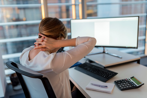 Rear View of a Tired Businesswoman Holding her Nape with Two Hands. Businesswoman feeling pain in neck after sitting at the table with desktop PC. Tired female suffering of office syndrome because of long hours computer work. Girl massaging her tense neck muscles.