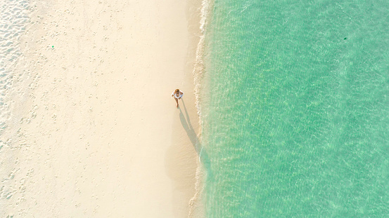 Aerial view of walking young woman on the sandy beach near sea with waves at sunset. Summer vacation in Lefkada island, Greece. Top view of sporty girl, azure water, mountain. Lifestyle and travel