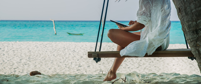 Low section shot of a woman using a digital tablet while sitting on a swing on a tropical beach,Maldives