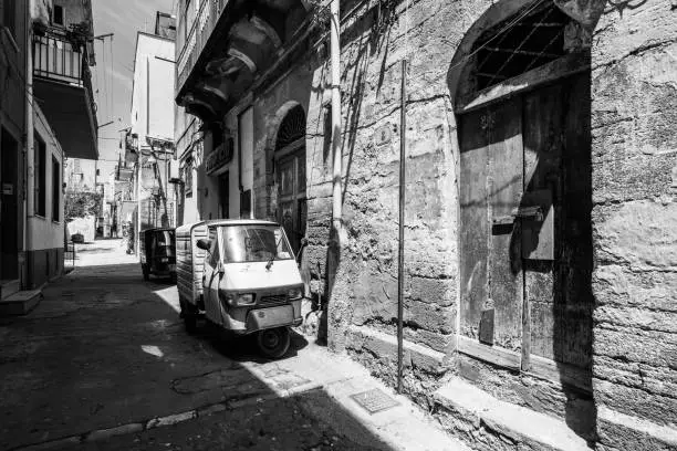 Black and white image of Sciacca old town with a Piaggio Ape three-wheeled vehicle near old buildings. Sciacca, Sicily, Italy, May 2018