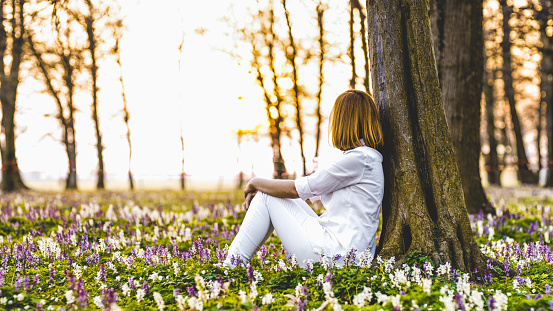 Side view shot of a woman in white clothing sitting among flowers under a tree in a forest