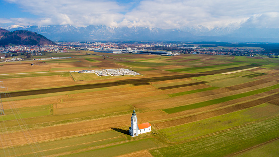 Aerial view of lone countryside church standing in patchwork field with city and mountain range in background,