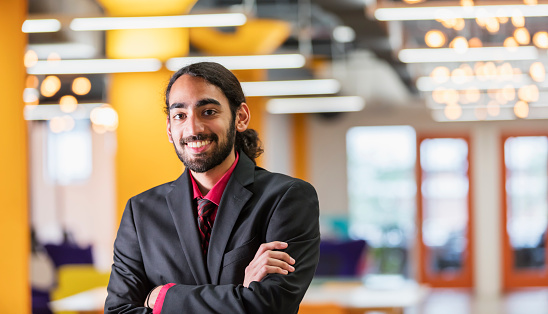 A young mixed race Indian and Caucasian man, in his 20s, with black hair, beard and mustache. He is a businessman wearing a suit and tie, looking at the camera, smiling with his arms crossed.