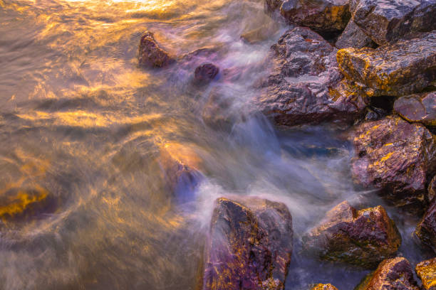 Long Exposure of waves washing over lakeshore rocks, illuminated by the golden light of sunrise.