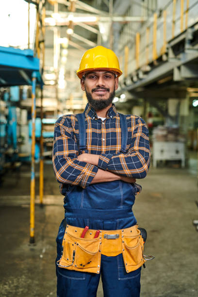 istock Smiling confident repair engineer with beard and mustache wearing hardhat and tool belt standing in factory shop and looking at camera 1094666446