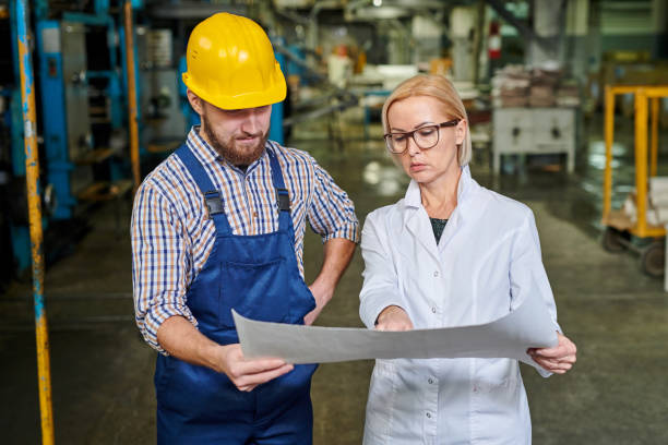istock Serious strict lady in lab coat pointing at blueprint while explaining sketch to brutal worker in industry 1094668266
