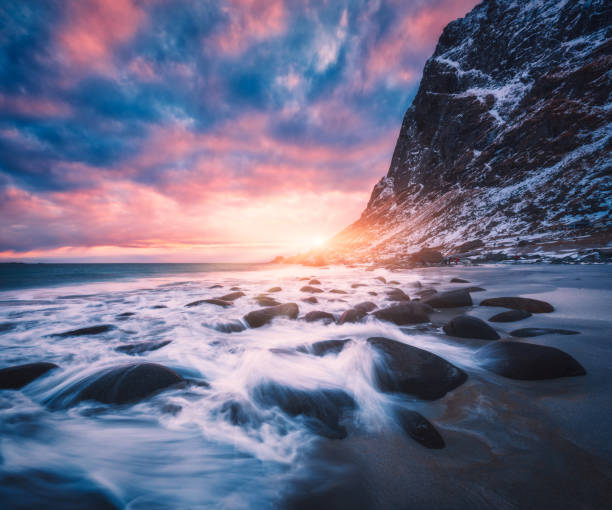 sandstrand mit steinen verschwommen wasser, blauer himmel mit rosa wolken und schneebedeckten berge bei sonnenuntergang. utakleiv beach, lofoten inseln, norwegen. winterliche landschaft mit meer, wellen, felsen, am abend - mountain peak norway reflection sunlight stock-fotos und bilder