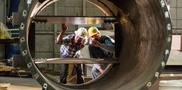 Two multi-ethnic workers in a metal fabrication factory inspecting a product, examining it with a flashlight. The mature man on the right is in his 40s and his African-American coworker is in his 30s.