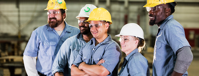 A group of five multi-ethnic workers wearing hardhats and safety glasses, standing in a dark warehouse. The team includes a mature woman in her 40s. They are standing side by side, facing the same directions, smiling.