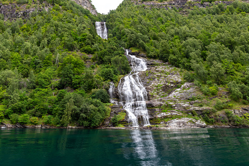 Trees and a waterfall are captured in the Geirangerfjord on a sunny summer day. The waterfall is flowing down the rocky mountain slope.