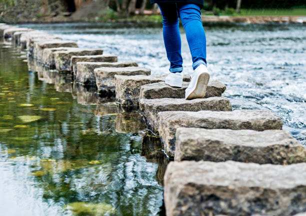 femme traversant les pierres de progression sur une rivière - stepping stone stone stepping footpath photos et images de collection