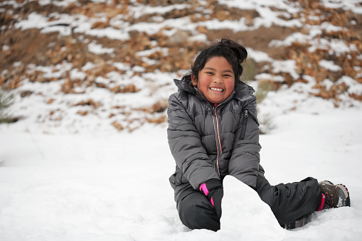 Smiling Latino girl sitting on snow covered mountain, making a snowman and wearing winter clothes.