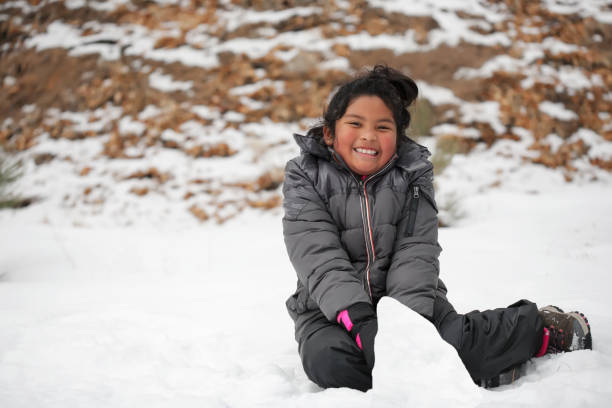 Smiling Latino girl sitting on snow covered mountain, making a snowman and wearing winter clothes. stock photo