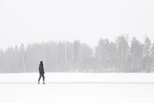 Young man alone slowly walking on snow covered roadside along forest in winter day. Side view.
