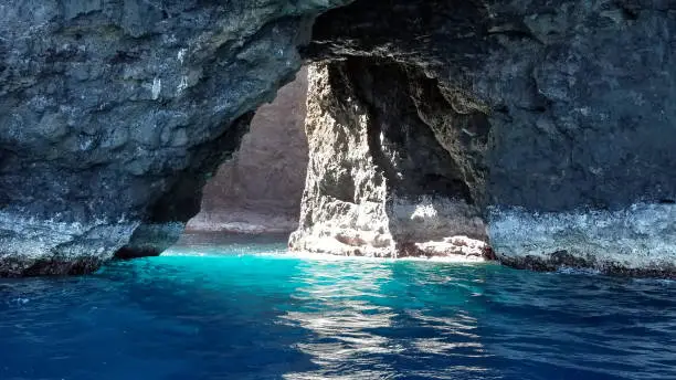 Entrance to a sea cave off of the Napali Coast on Kauai, Hawaii that shows different hues of blue in the water as the natural sun light pierces through a hole up above in the cave.