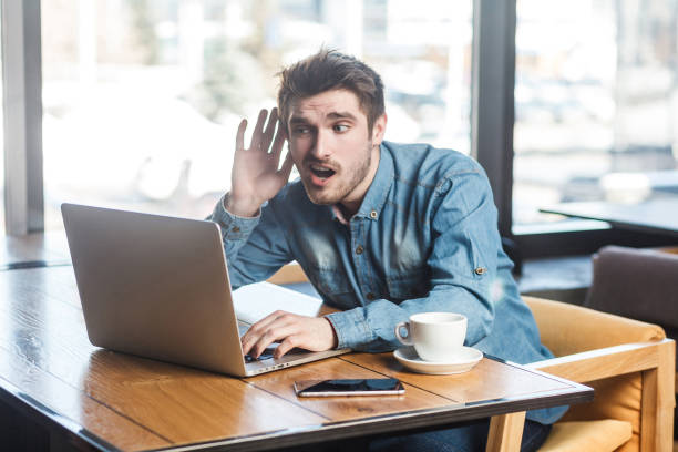 retrato do modo de exibição lado interessante de barbudo jovem freelancer em camisa jeans azul estão sentados no café e fazer chamada de vídeo no laptop, se inclinar a mão na orelha que não ouvir a companheira. - spy secrecy top secret mystery - fotografias e filmes do acervo