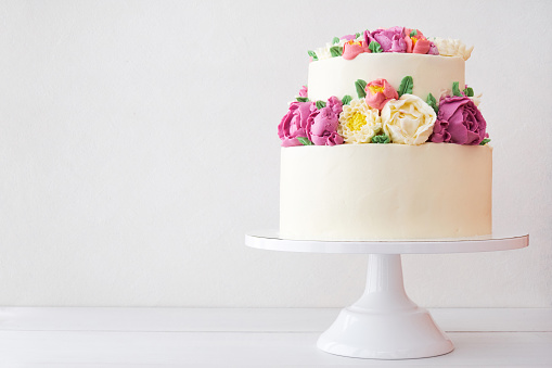 Two-tiered white wedding cake decorated with color cream flowers on a white wooden background.