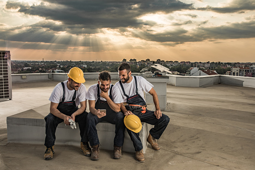 Young happy construction workers relaxing on a rooftop and reading text message on cell phone.