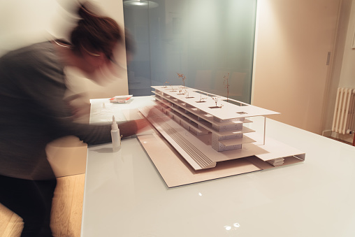 Female architect working on architecture model made with cardboard on table in office of architectural firm. She is moving, motion blurred.
