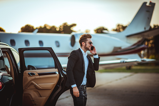 Young rich manager talking on a smart phone while getting out of a luxurious car parked next to a private airplane on a taxiway.