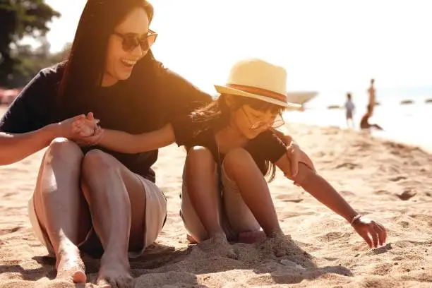 Photo of Asian girl wearing a hat and sunglasses and his mother sitting on the sand at the beach