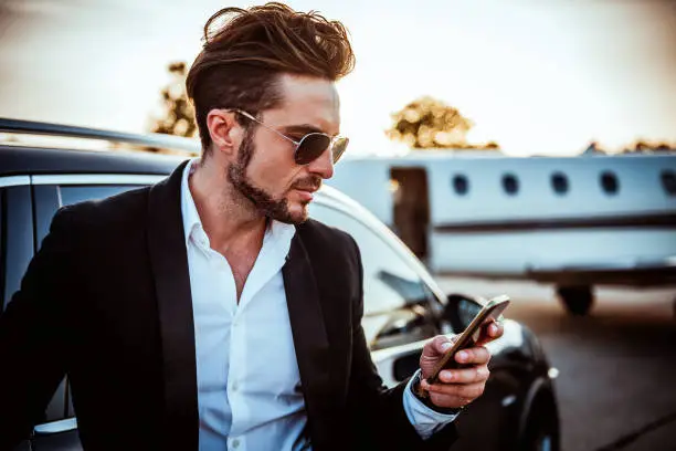 Photo of Rich entrepreneur using a mobile phone with a luxurious black car and a private jet parked behind him