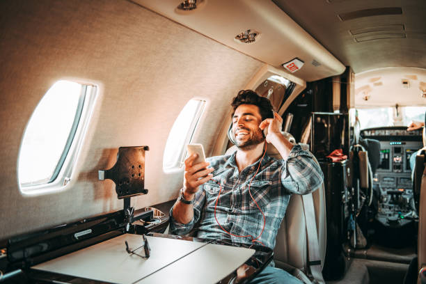 joven rico hombre escuchando música por los auriculares y el uso de un teléfono móvil mientras estaba sentado en un avión privado - status symbol audio fotografías e imágenes de stock